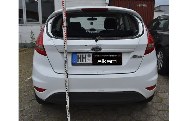 Heckansicht eines weißen Ford Fiesta mit eingeschlagener Heckscheibe und Messlatte zur Schadensdokumentation.